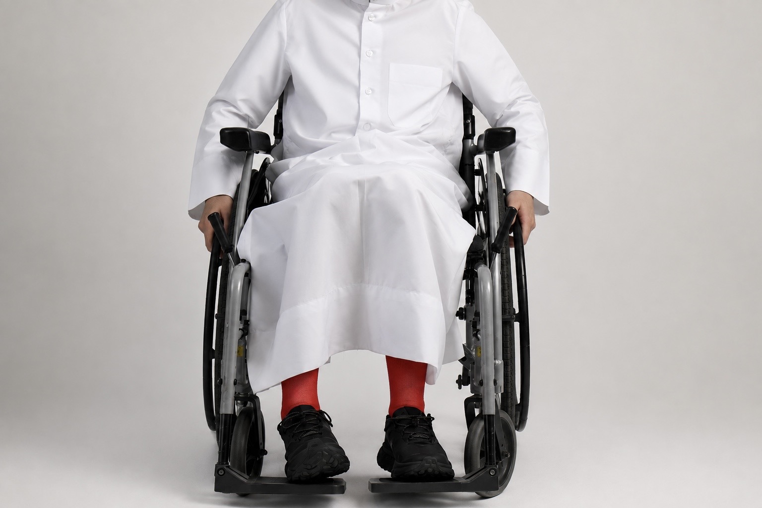 A person in a white thobe and bright red socks seated in a manual wheelchair, hands resting on the wheel rims, against a plain light gray backdrop.
